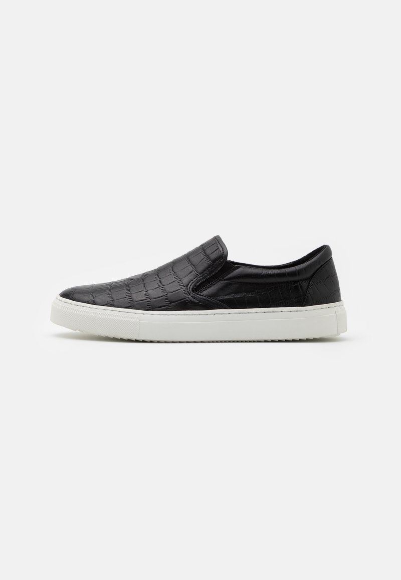 Baskets noires sans lacets avec un motif texturé et une épaisse semelle en caoutchouc blanche, vues de côté sur un fond clair uni.