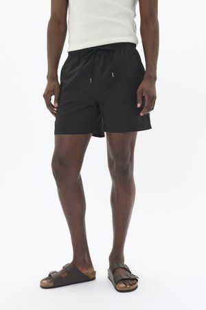 Schwarze Schwimmshorts aus leichtem Stoff mit elastischem Bund und Kordelzug. Kombiniert mit braunen Riemchensandalen.