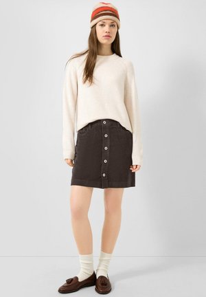 Femme portant un pull crème, une jupe en velours côtelé foncée boutonnée, un bonnet rayé, des chaussettes blanches et des mocassins marron à pompons, debout devant un fond uni.