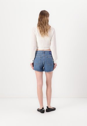 Mujer con cabello largo y ondulado que lleva un suéter tejido blanco corto, shorts de mezclilla azul de talle alto y zapatos negros sin cordones, de pie contra una pared blanca.