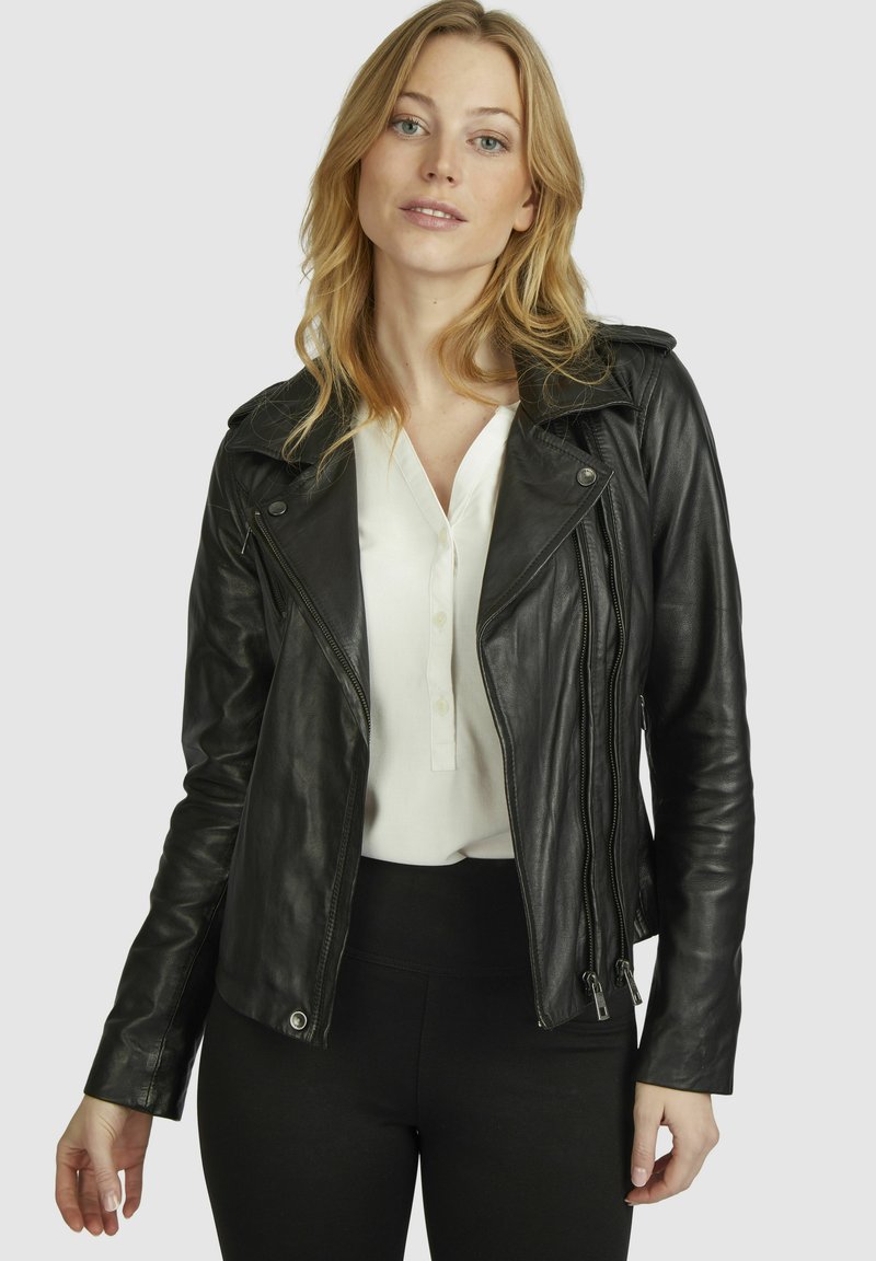 Schwarze Lederbikerjacke mit Reißverschlüssen, einreihigem Kragen und taillierter Silhouette, getragen über einem weißen Hemd mit Knöpfen und schwarzen Leggings.