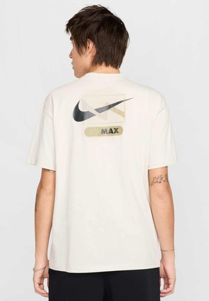 T-shirt Nike Air Max in bianco sporco, con un grande design del logo nero e oro sulla schiena. Maniche corte, vestibilità rilassata, materiale in cotone morbido.