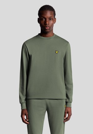 Jonge man draagt een bijpassend olijfgroen shirt met lange mouwen en broek, met een klein geel vogel-logo op de borst en dij, tegen een grijze achtergrond.