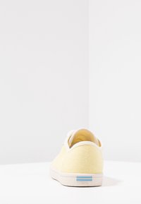 Baskets en toile jaunes avec une surface texturée, accents blancs et un logo à rayures bleues sur le talon. Forme arrondie, design minimaliste.