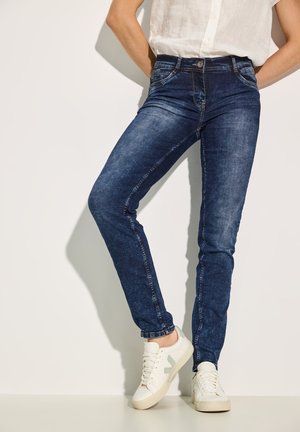 Sininen farkku skinny-farkut, joissa on haalistunut pinta, viisi taskua ja etulukko, yhdistettynä valkoisiin tennareihin, joissa on vihreitä yksityiskohtia.