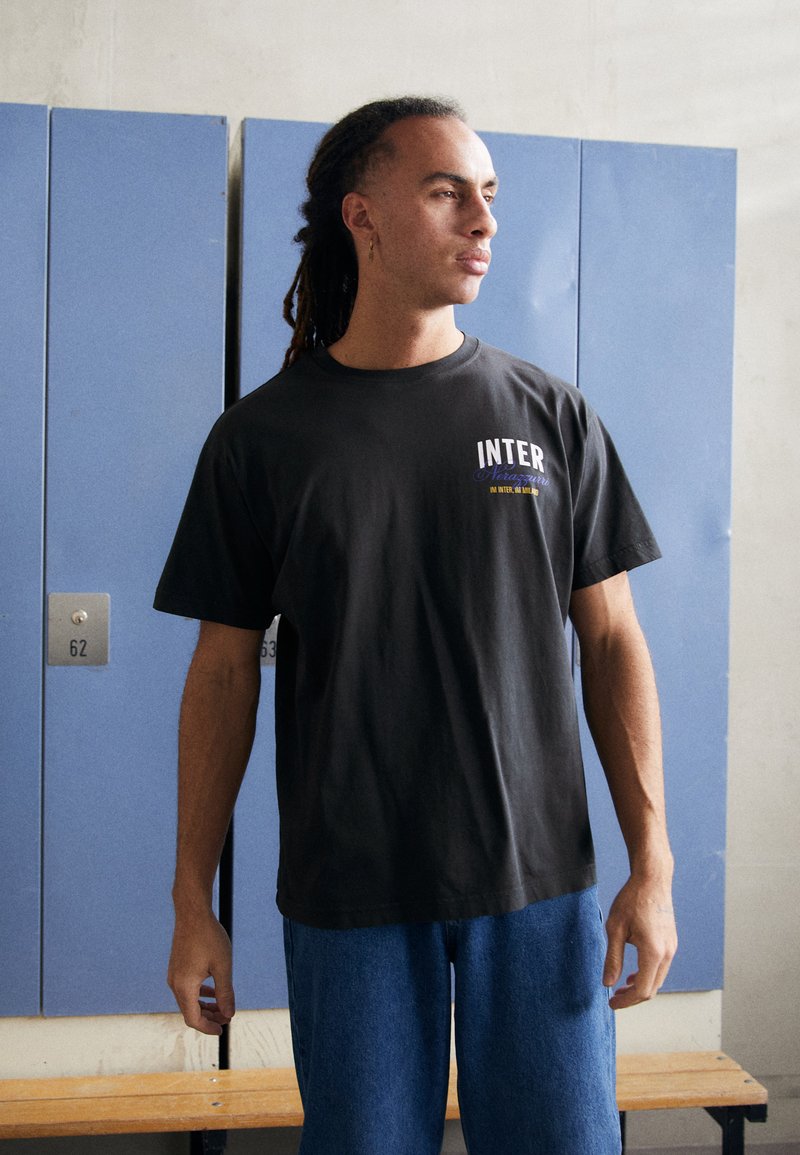 Schwarzes Baumwoll-T-Shirt mit kurzen Ärmeln, das das "INTER"-Logo in Weiß und Lila zeigt, kombiniert mit blauen Jeans. Einfacher, lässiger Schnitt.