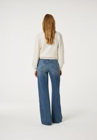 Lichtbeige gebreide trui met pofmouwen, gecombineerd met een paar wijdvallende blauwe denim jeans met zijknopen en een vervaagde wassing, gezien van achteren.