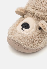 Chausson en peluche beige avec un extérieur texturé et doux. Dispose d'un œil rond noir cousu et de petites oreilles, avec une semelle beige clair.