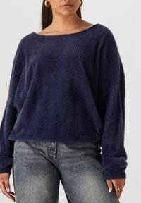 Marineblauwe, fuzzy pullover trui met een off-the-shoulder halslijn en lange mouwen, gecombineerd met licht versleten grijze jeans.