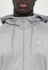 Lys grå zip-up hættetrøje med snoretræk i hætten. Har et lille EA7-logo patch på brystet og et glat, tekstureret stof.