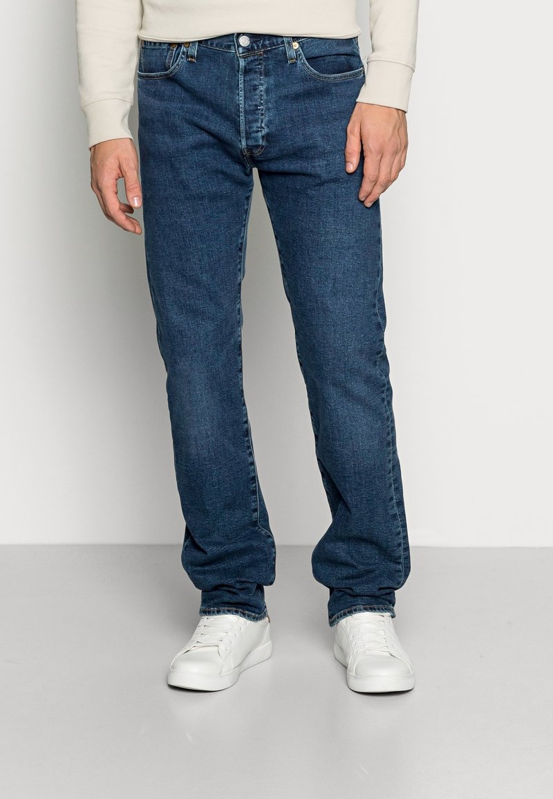 Homme portant un jean droit bleu foncé et des baskets blanches, debout devant un fond clair uni.