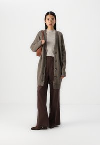 Cardigan lung, cu deschidere frontală, dintr-un material moale și pufos, de culoare gri-maro, cu butoane mari, purtat peste un top bej cu dungi și pantaloni lejeri maro cu talie înaltă.