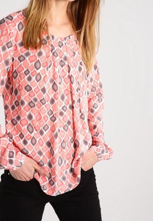 Nainen pukeutuneena pitkähihainen pusero, jossa on vaaleanpunainen, valkoinen ja harmaa abstrakti kuvio, kädet mustien housujen taskuissa, yksiväristä taustaa vasten.