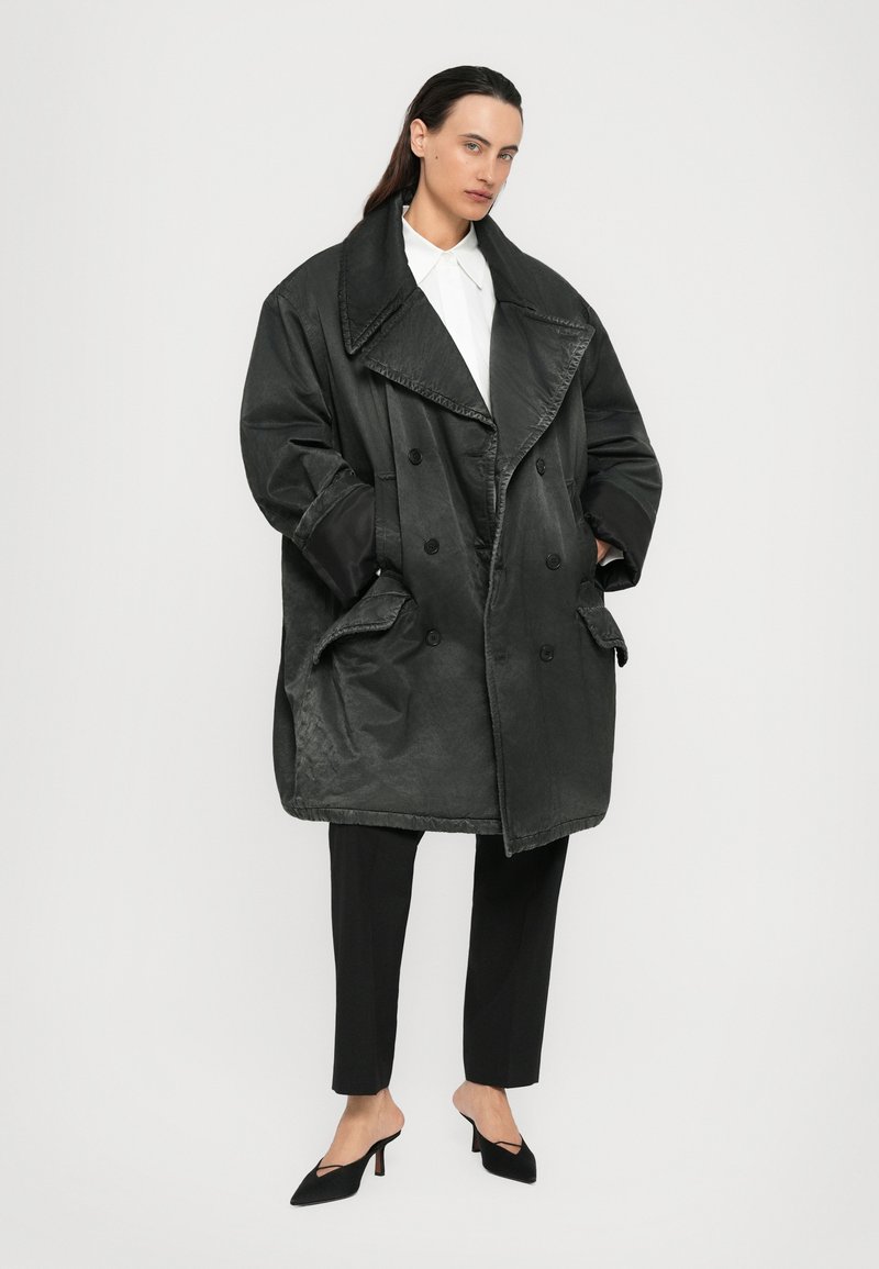 Kvinna som bär en överdimensionerad svart dubbelfäppad kappa, svarta byxor, vit skjorta och svarta slingback-stövlar, stående mot en neutral bakgrund.