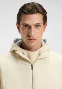 Giacca beige con zip e cappuccio, caratterizzata da una texture liscia e un rivestimento interno grigio. Indossata sopra un top di colore chiaro.