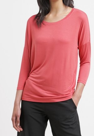 Kobieta ubrana w koralowo-różową bluzkę z długim rękawem i dekoltem w łódkę oraz czarne spodnie, stojąca z jedną ręką w kieszeni na jednolitym tle.