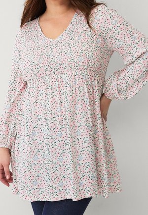 Woman wearing a long-sleeve, pink floral patterned blouse with a V-neck and gathered detailing around the chest.