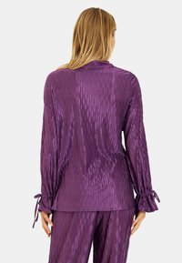 Top de satén púrpura con mangas largas, que presenta patrones texturados en forma de ondas y puños anudados, combinado con pantalones a juego.