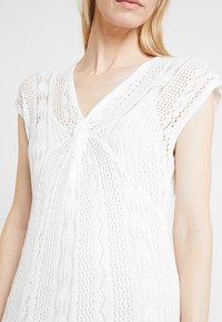 Top bianco lavorato a maglia con scollo a V, maniche corte e motivi testurizzati con un dettaglio centrale attorcigliato, realizzato in un filato leggero.
