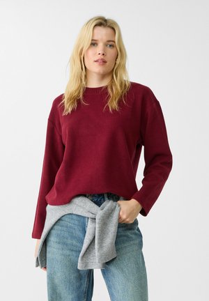 Pull bordeaux avec un col rond et des manches longues, associé à un jean bleu clair et un pull gris noué à la taille. Texture lisse.