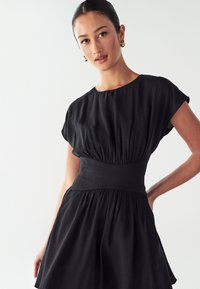 Woman wearing a black short-sleeve dress with wide waistband and gathered skirt, posing with one hand on her hip against plain background.