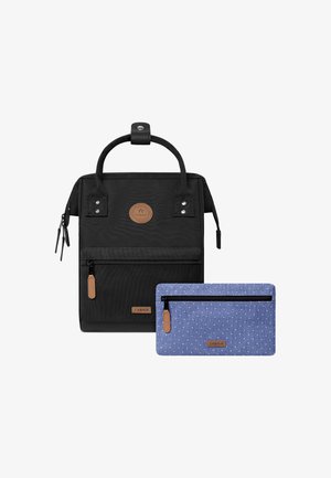 Sac à dos en tissu noir au fini texturé, arborant un logo en cuir marron, une poche zippée à l'avant et une pochette à pois bleus en dessous.