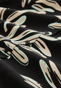 Zwarte stof met een patroon van abstracte beige en mintgroene vormen. Twee zwarte knopen zijn zichtbaar en voegen een textuurdetail toe.