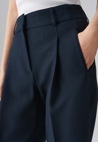 Pantalons navy avec un devant plissé, des poches latérales et une texture lisse. Fabriqués à partir d'un tissu structuré, avec une coupe ajustée.