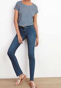 Graues T-Shirt mit kurzen Ärmeln und Strassdetails auf der Vorderseite, kombiniert mit dunkelblauen Skinny Jeans und metallisch roségoldenen Sandalen.