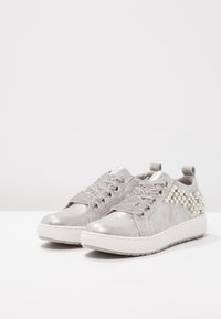 Srebrne sneakersy z satynowymi sznurowadłami i perłowymi ozdobami na pięcie, z grubymi białymi gumowymi podeszwami na białej powierzchni.