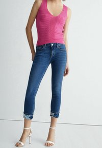 Mujer vistiendo una blusa sin mangas de punto rosa, jeans skiny azules con dobladillo y estampado floral, y sandalias de tacón blanco, de pie contra un fondo liso.