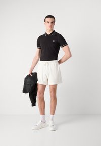 Fekete poloing fehér kiegészítőkkel, fehér sorttal párosítva. A modell egy fekete dzsekit tart, fehér sportcipőt és zoknit visel.