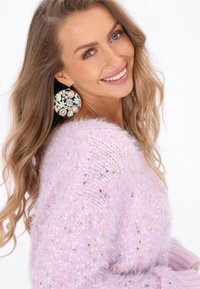 Flauschiger rosa Pullover mit mehrfarbigen Sprenkeln. Große runde Ohrringe mit floralen Mustern in Pastellfarben und metallischen Akzenten. Neutraler Hintergrund.