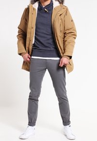 Hnědá parka s kožešinovou kapucí, tmavě šedá mikina pod ní, šedé kalhoty a bílé tenisky. Materiál je hladký s volným střihem.