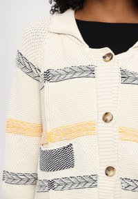 Kremowy kardigan z teksturowanym wzorem dzianiny, ozdobiony niebieskimi, czarnymi i żółtymi paskami. Zawiera dwa guziki oraz detal z kieszonką.
