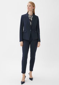 Marineblauw op maat gemaakt pak met een getailleerde blazer met een enkele knoop, gecombineerd met slim fit broek en puntige hakken.