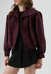Bordeauxrode, zachte stoffen jas met een kraag, knoopsluiting en pofmouwen. Draagt over een zwarte top en gecombineerd met een zwarte rok.