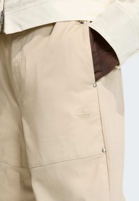 Béžové sportovní kalhoty s hladkým povrchem, bočními kapsami a reliéfním logem. Obsahují stříbrné patentky a detaily šití.