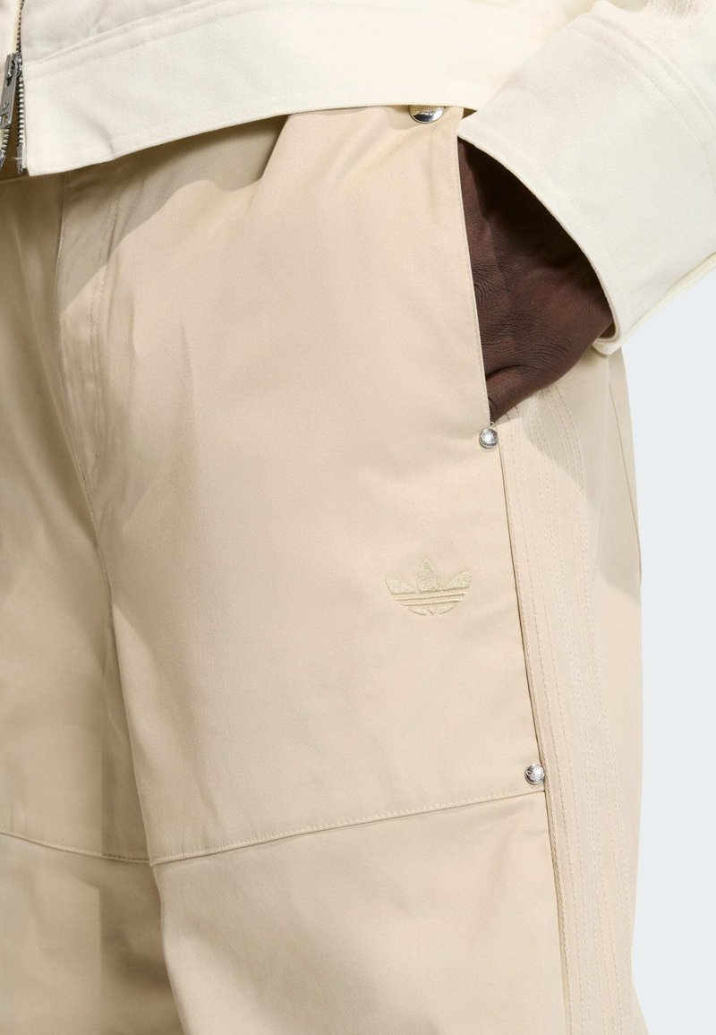 Beige athletic pants featuring a smooth texture, side pockets, and an embossed logo. Includes silver snap buttons and stitching details.