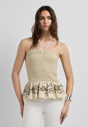 Femme portant un haut beige smocké à col licou avec un ourlet à volants et une broderie florale foncée, associé à un pantalon blanc et un bracelet manchette argenté.