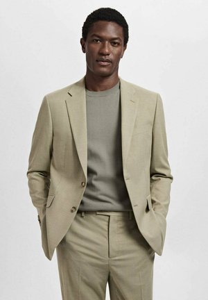 Light beige suit with a structured jacket, matching trousers, and a gray knit sweater. Smooth fabric, tailored fit, and gold-tone buttons.