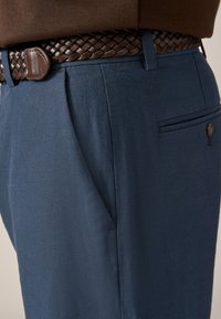 Gros plan sur un pantalon bleu marine avec poches latérales et arrière, porté avec une ceinture en cuir tressé marron foncé et un haut sombre.