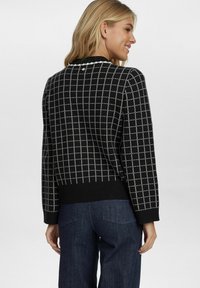 Un pull noir avec un motif à carreaux blancs, un ourlet et des poignets côtelés, et un détail de bouton au dos, porté avec un jean bleu foncé.