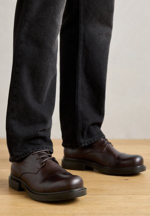 Braune Lederschuhe mit abgerundeter Spitze, Schnürdesign und dicker schwarzer Gummisohle, kombiniert mit dunkelblauen Jeans.