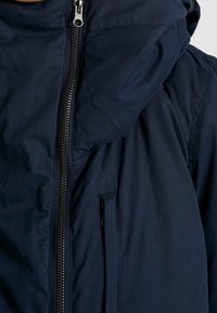 Zbliżenie na granatową kurtkę z przednim zamkiem błyskawicznym i pionową kieszenią zapinaną na zamek na wysokości klatki piersiowej.