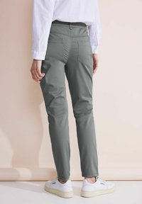 Personne portant un pantalon gris à jambe droite, une chemise blanche à manches longues et des baskets blanches, debout devant un fond neutre.