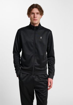 Zwarte sportjack met ritssluiting, gemaakt van gladde stof, met een hoge kraag en een klein wit logo op de linkerborst. Gecombineerd met bijpassende broek.