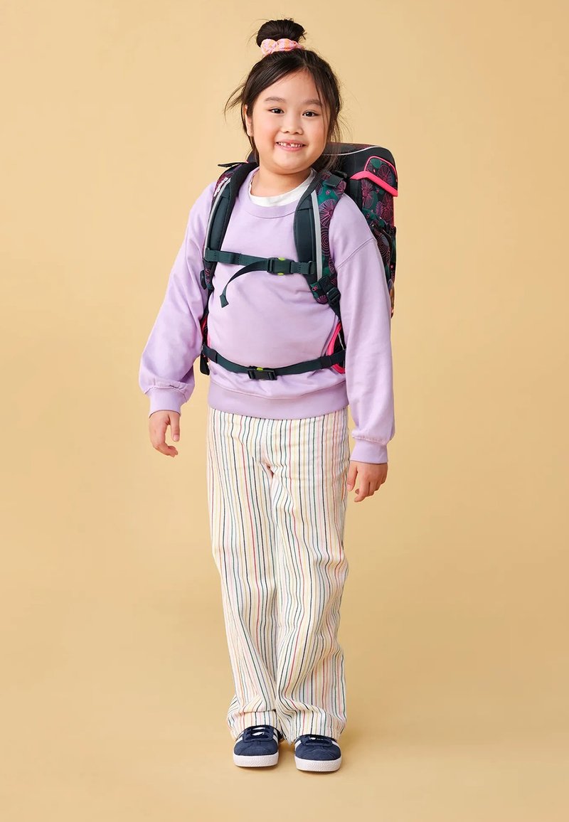 Purple sweatshirt, striped pants, and blue shoes. A colorful backpack with pink accents. The child has a bun hairstyle with a hair tie.