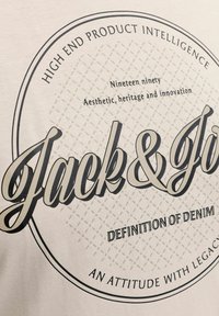 Krem t-shirt med et sort logo i midten med teksten "Jack & Jo" skrevet med kursiv, omgivet af cirkulær tekst og et baggrundsmønster af diamanter.