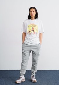 White T-shirt with a colorful graphic print, gray cargo sweatpants, and beige athletic shoes, standing on a blue carpet background.